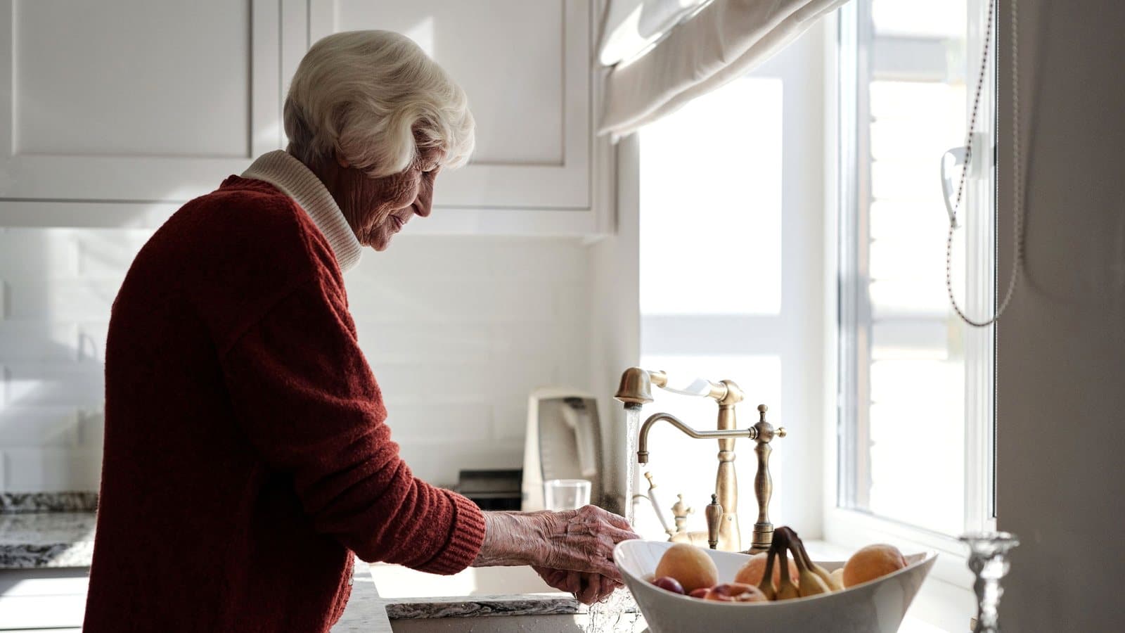 Ældre kvinde står ved køkkenvasken i sit eget hjem og skyller frugt i dagslyset fra vinduet