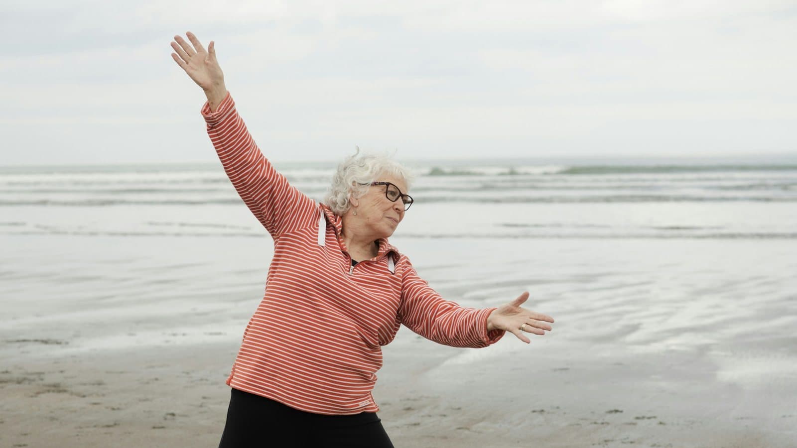 Ældre kvinde laver balanceøvelser med udstrakte arme på en strand