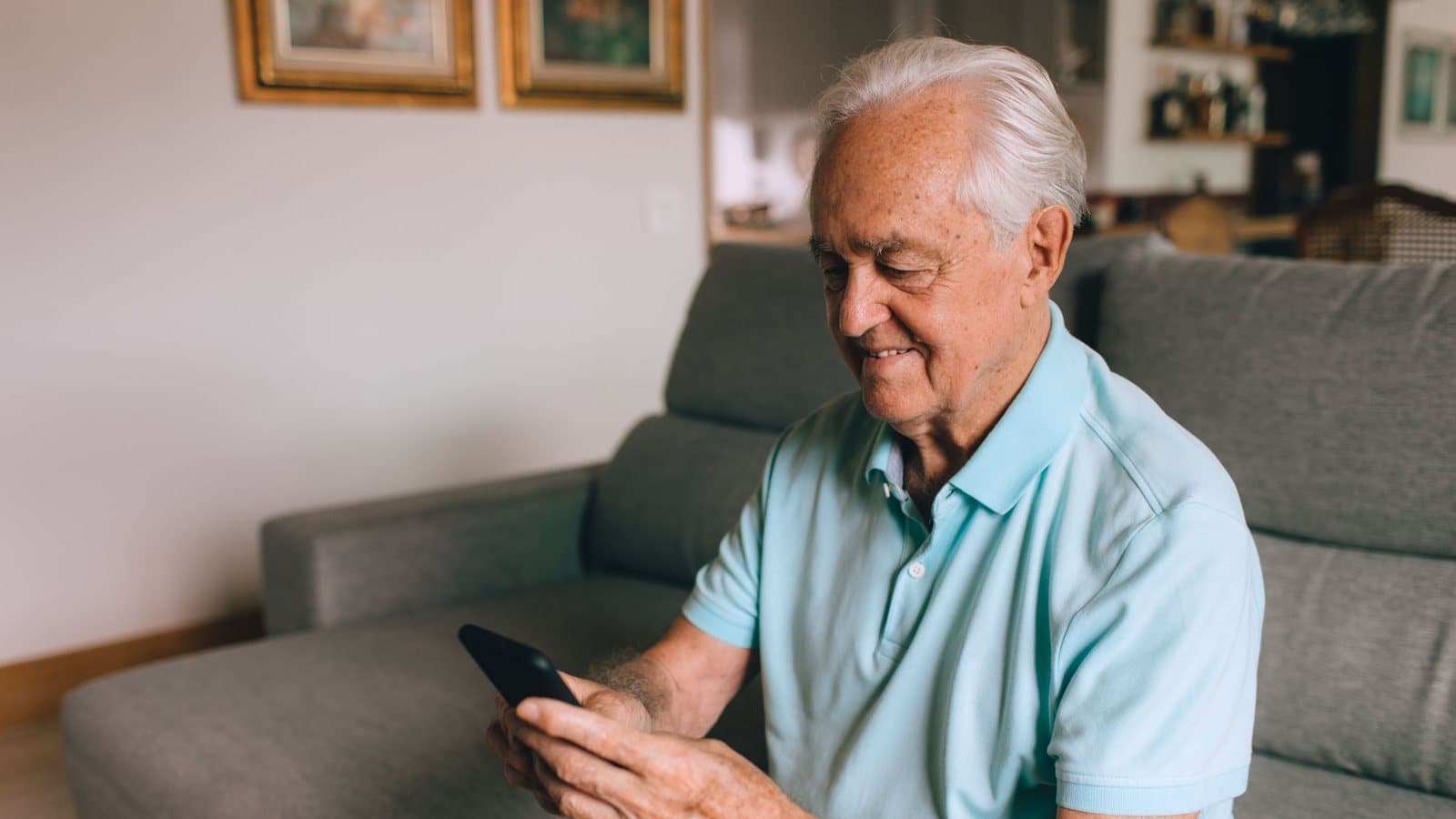 Ældre mand sidder i sofaen derhjemme og kigger smilende på sin smartphone